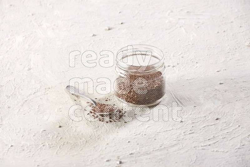 A glass jar full of flax seeds with a metal spoon full of the seeds on a textured white flooring