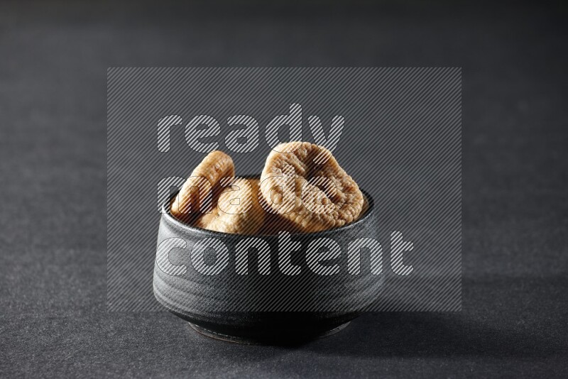 A black pottery bowl full of dried figs on a black background in different angles