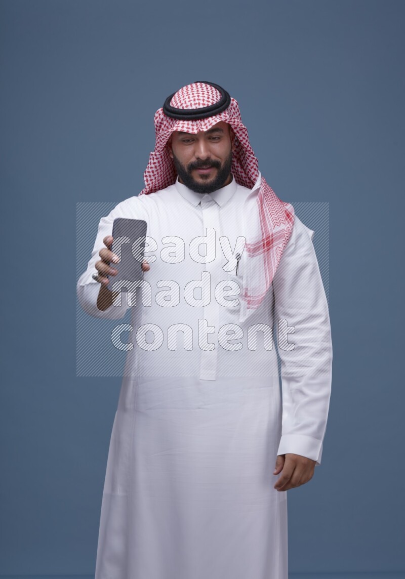 A man Showing his smartphone Screen on Blue Background wearing Saudi Thob