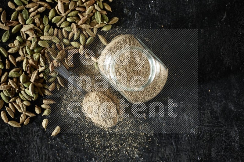 A glass spice jar and a metal spoon full of cardamom powder and cardamom seeds spreaded on textured black flooring
