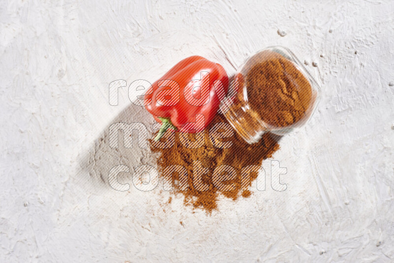 A glass jar full of ground paprika powder flipped with some spilling powder on white background