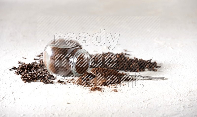 A flipped glass spice jar and a metal spoon full of cloves powder and powder came out of the jar with cloves spread on textured white flooring