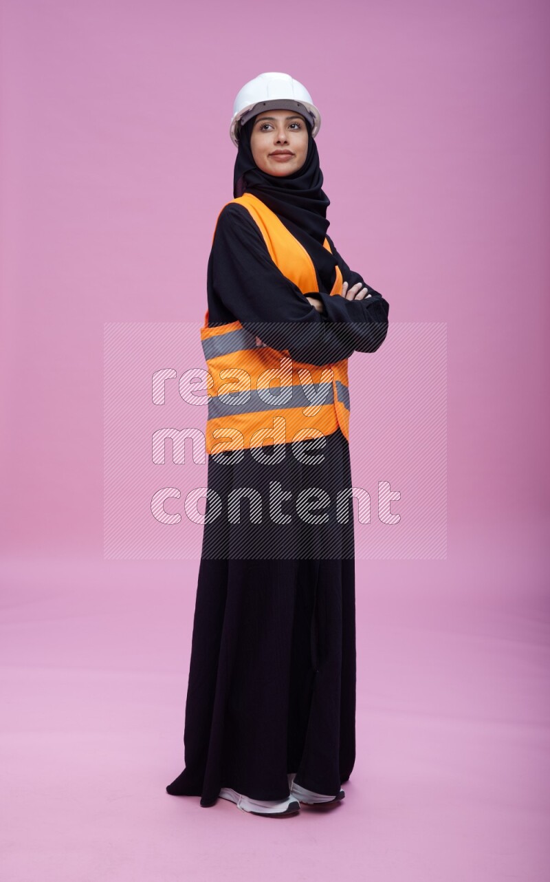 Saudi woman wearing Abaya with engineer vest and helmet standing with crossed arms on pink background