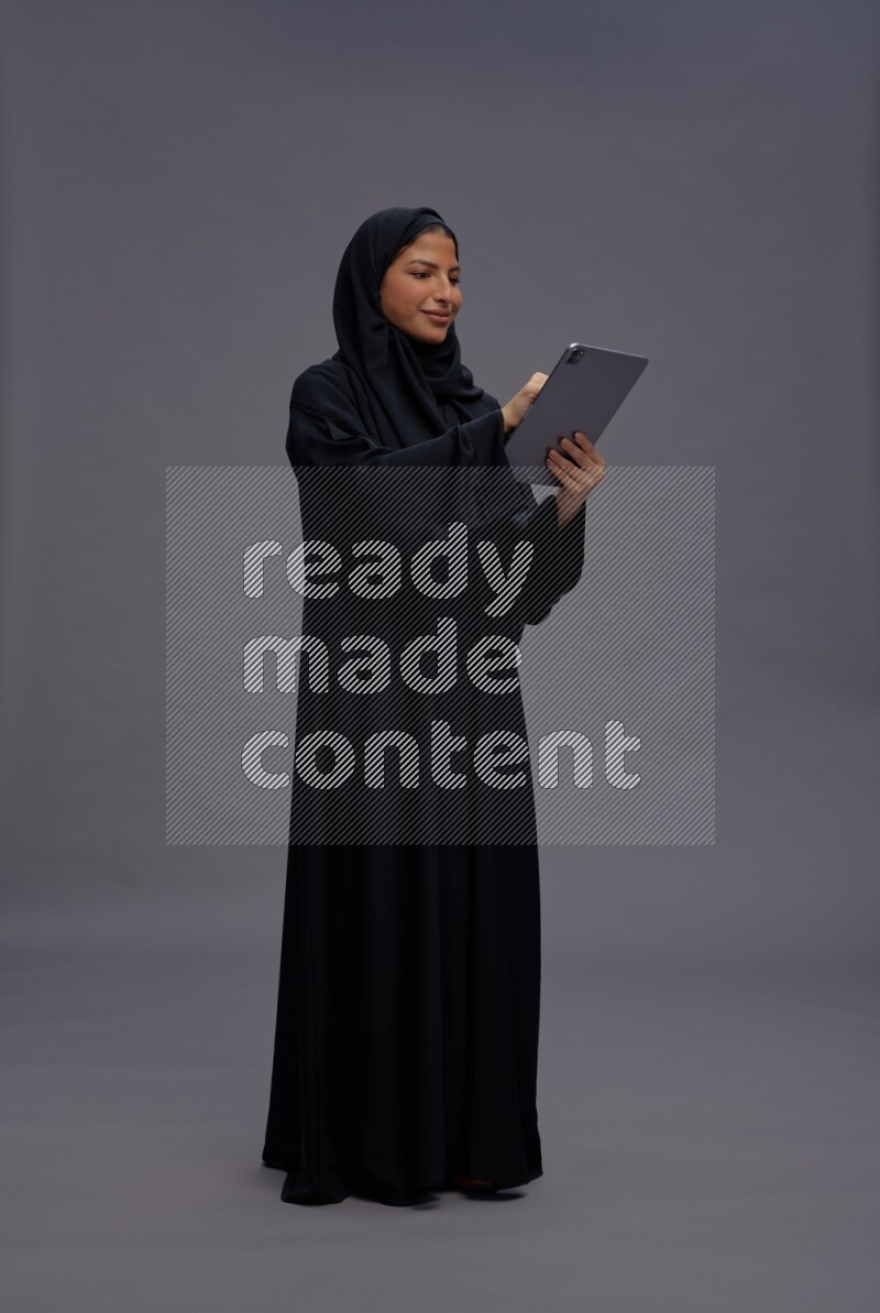 Saudi woman wearing Abaya standing working on tablet on gray background