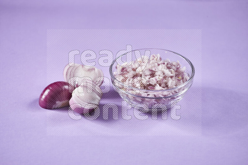 A glass bowl full of chopped red onions with halved onions beside it on purple background