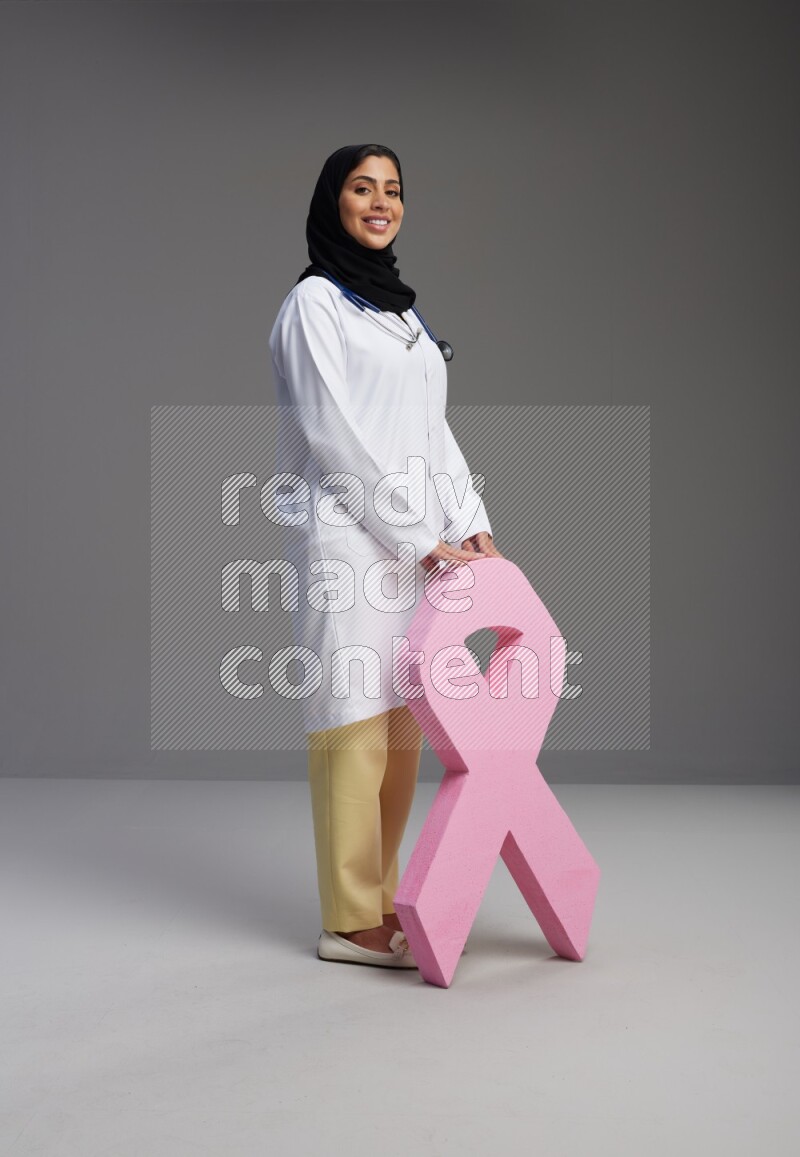 Saudi woman wearing lab coat with stethoscope standing holding awareness ribbon symbol standing on Gray background