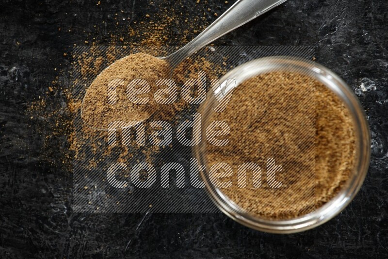 A glass jar and a metal spoon full of cumin powder on a textured black flooring