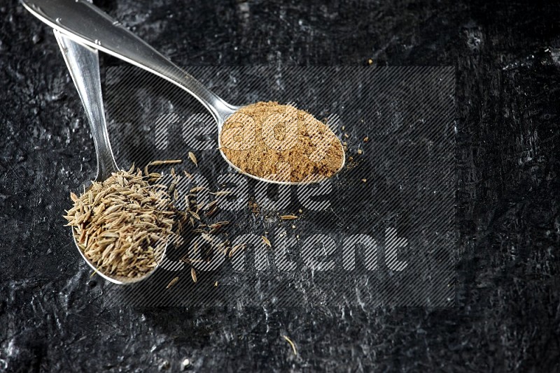 2 metal spoons full of cumin seeds and cumin powder on a textured black flooring