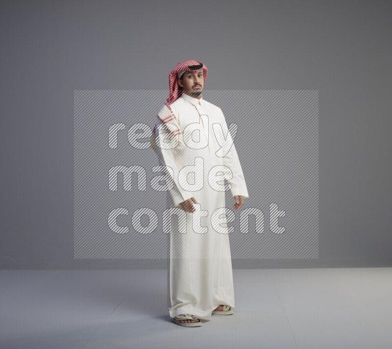 A Saudi man standing wearing thob and red shomag interacting with the camera on gray background