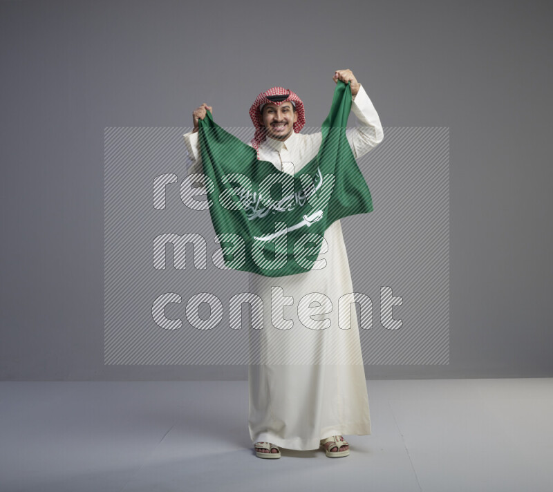 A saudi man standing wearing thob and red shomag holding big saudi flag on gray background