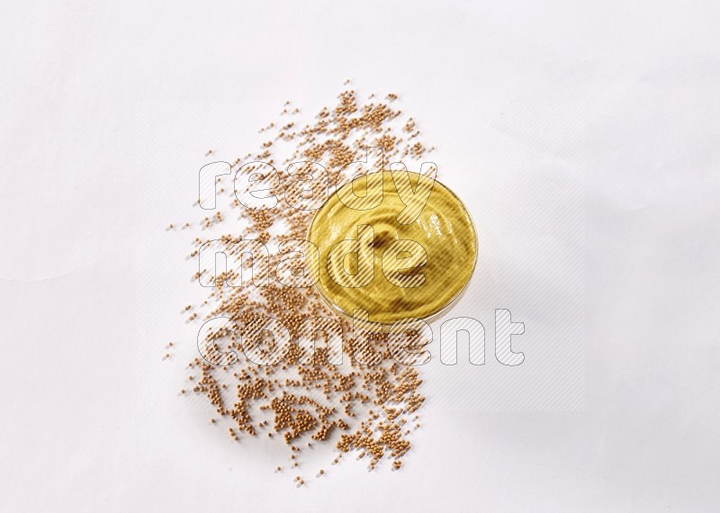 A glass bowl full of mustard paste with mustard seeds underneath on white flooring in different angles