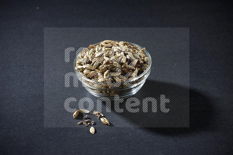 A glass bowl full of cardamom and more seeds spreaded beneath the bowl on black flooring