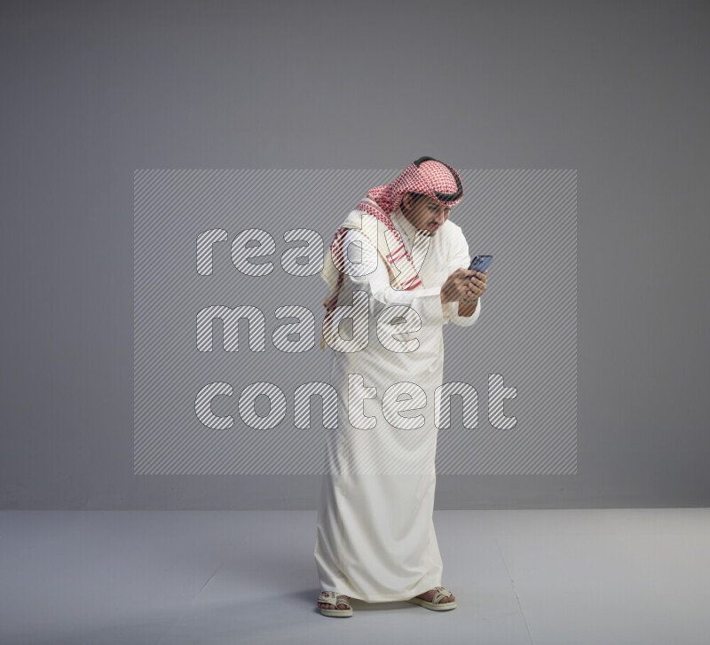 A  Saudi man standing wearing thob and red shomag texting on phone on gray background