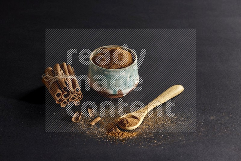 Ceramic bowl full of cinnamon powder and a wooden spoon full of powder with cinnamon sticks stacked and bounded on black background