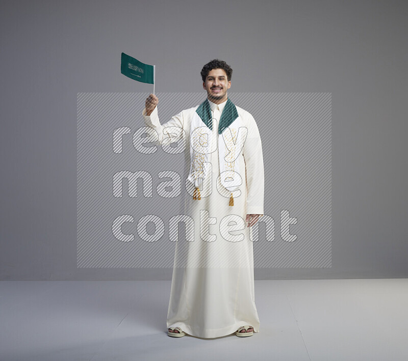 A Saudi man standing wearing thob and saudi flag scarf and holding small saudi flag on gray background