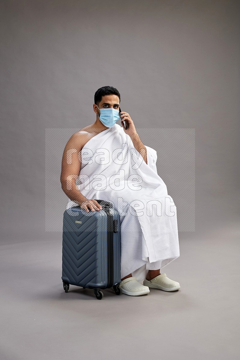 A man wearing Ehram sitting on chair holding traveling bag on gray background