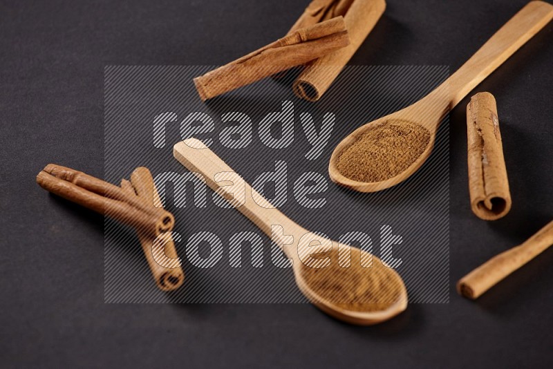 Two wooden spoons full of cinnamon powder with cinnamon sticks on black background