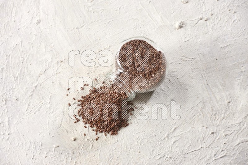 A glass spice jar full of flax seeds flipped and seeds spread out on a textured white flooring