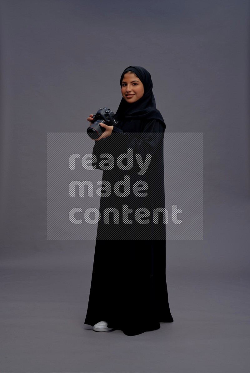 Saudi woman wearing Abaya standing holding Camera on gray background