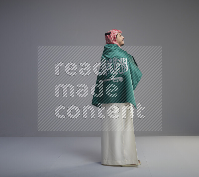A Saudi man standing wearing thob and red shomag wrapping big saudi flag on gray background