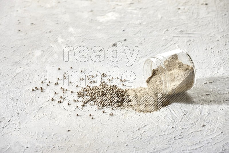 A flipped glass jar full of white pepper powder with spilled powder and white pepper beads on textured white flooring