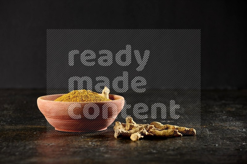 A wooden bowl full of turmeric powder with dried turmeric whole fingers on textured black flooring