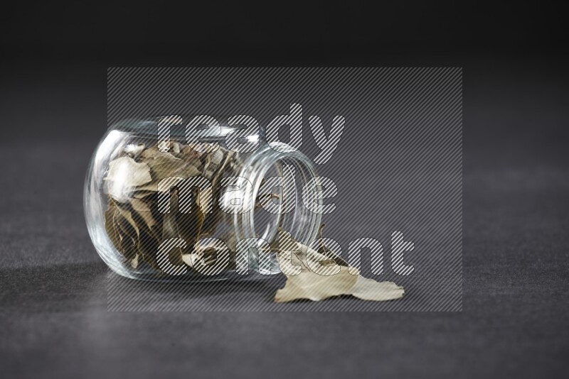 A glass spice jar full of dried bay leaves and jar is flipped and leaves spread out on black flooring