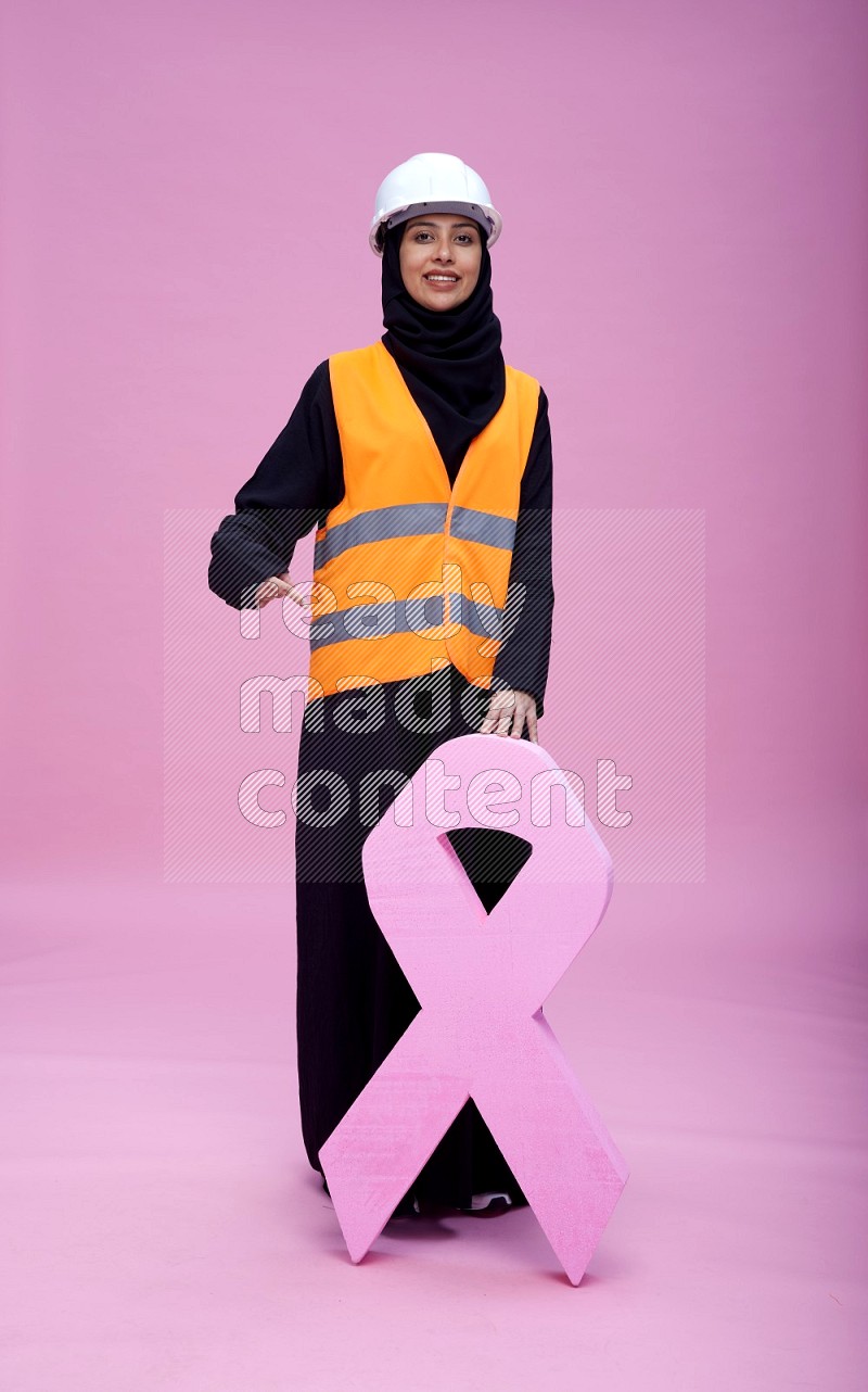 Saudi woman wearing Abaya with engineer vest and helmet standing awareness ribbon on pink background