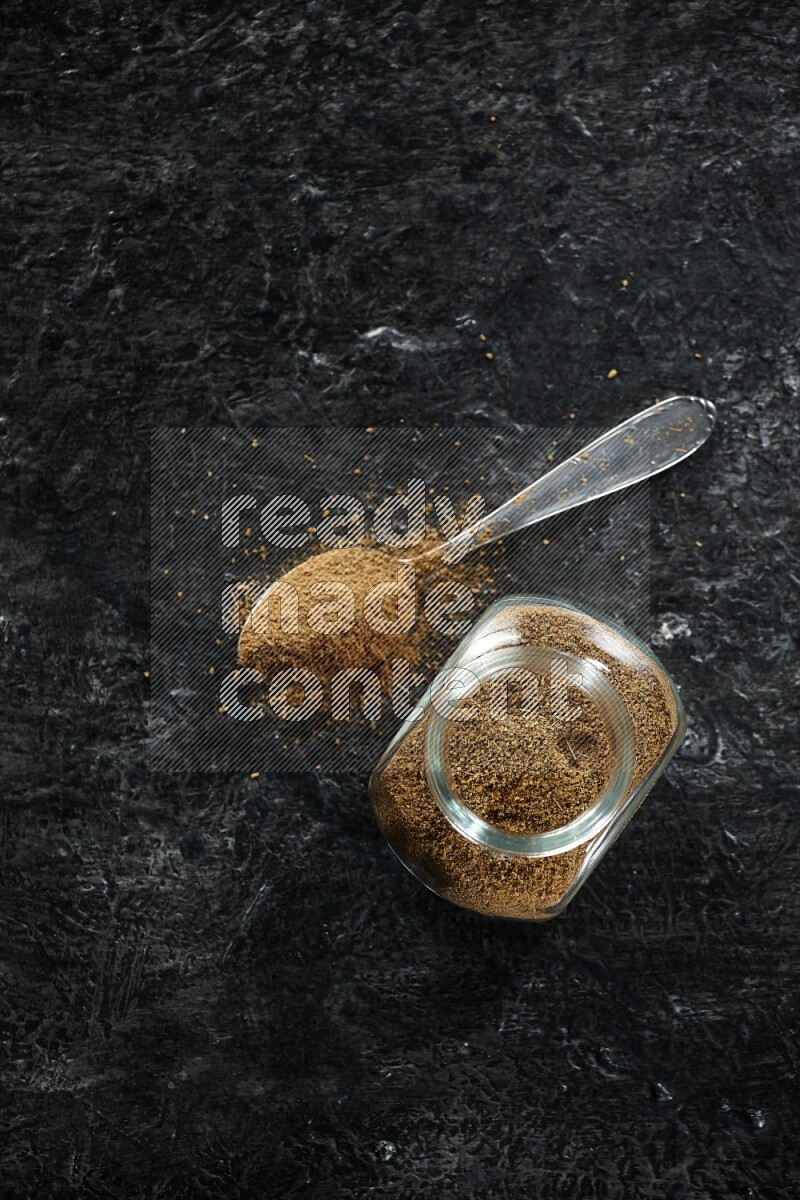 A glass spice jar and a metal spoon full of cumin powder on a textured black flooring