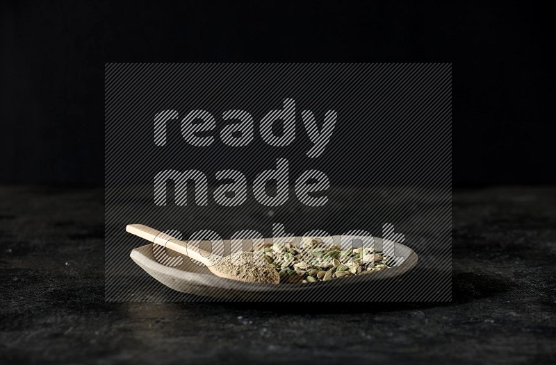 A plate filled with cardamom seeds and a wooden spoon full of cardamom powder on a textured black flooring