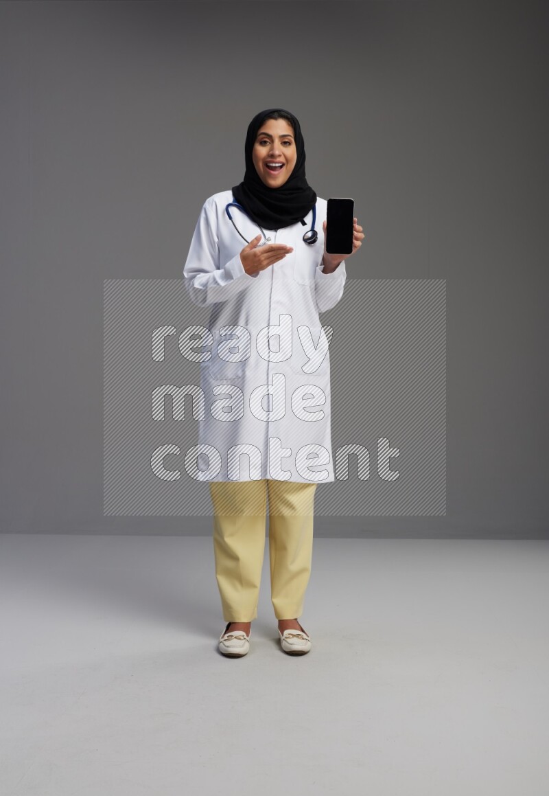 Saudi woman wearing lab coat with stethoscope standing showing phone to camera with sign in the back on Gray background