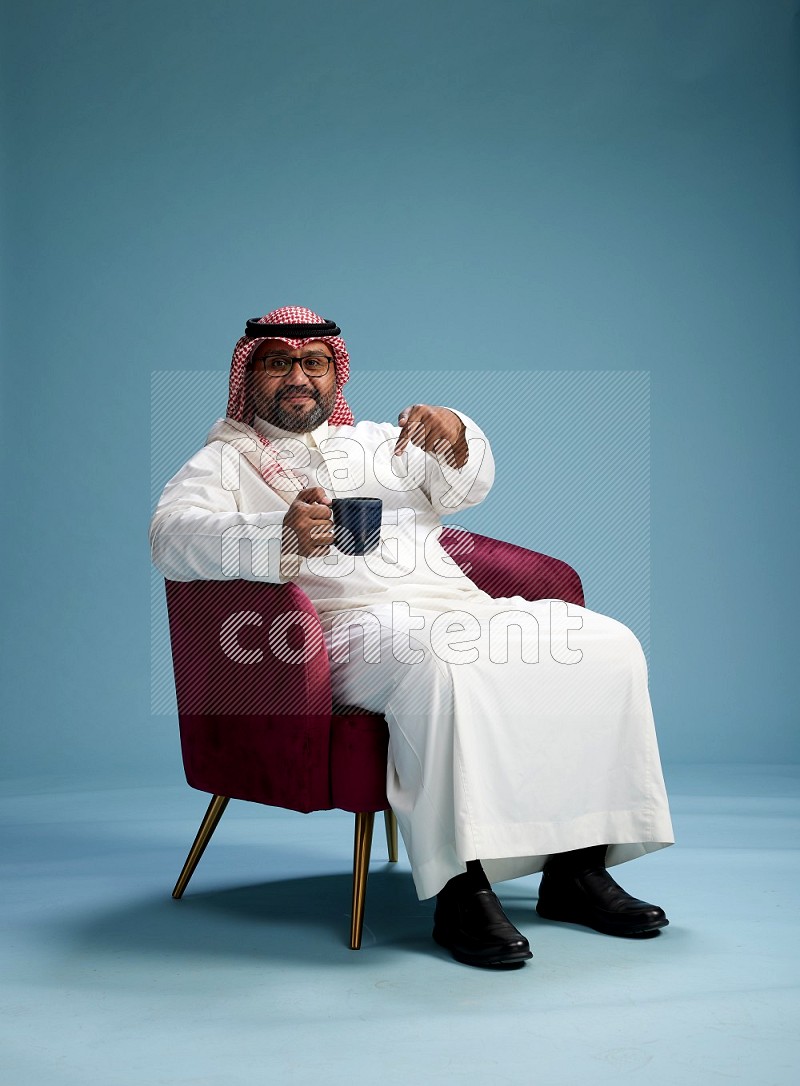 Saudi Man with shimag sitting on chair drinking coffee on blue background