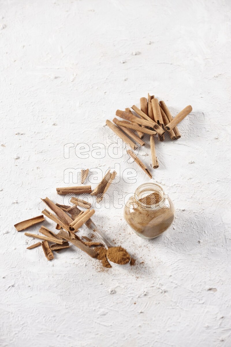 Herbal glass jar full cinnamon powder and a metal spoon surrounded by cinnamon sticks on a white background
