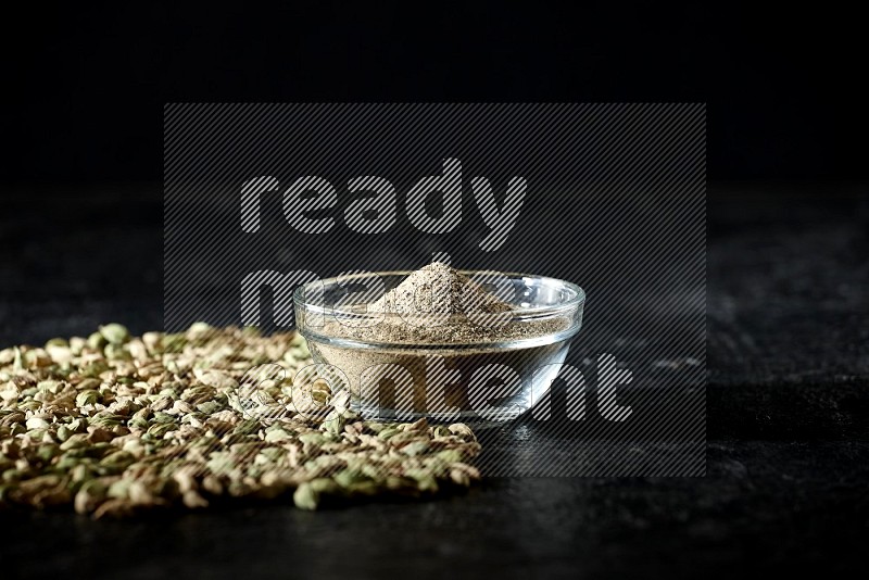 A glass bowl full of cardamom powder and cardamom seeds spreaded beneath the bowl on textured black flooring