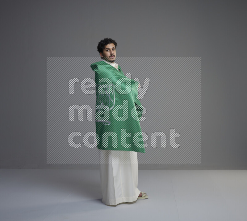 A Saudi man standing wearing thob wrapping big Saudi flag on gray background