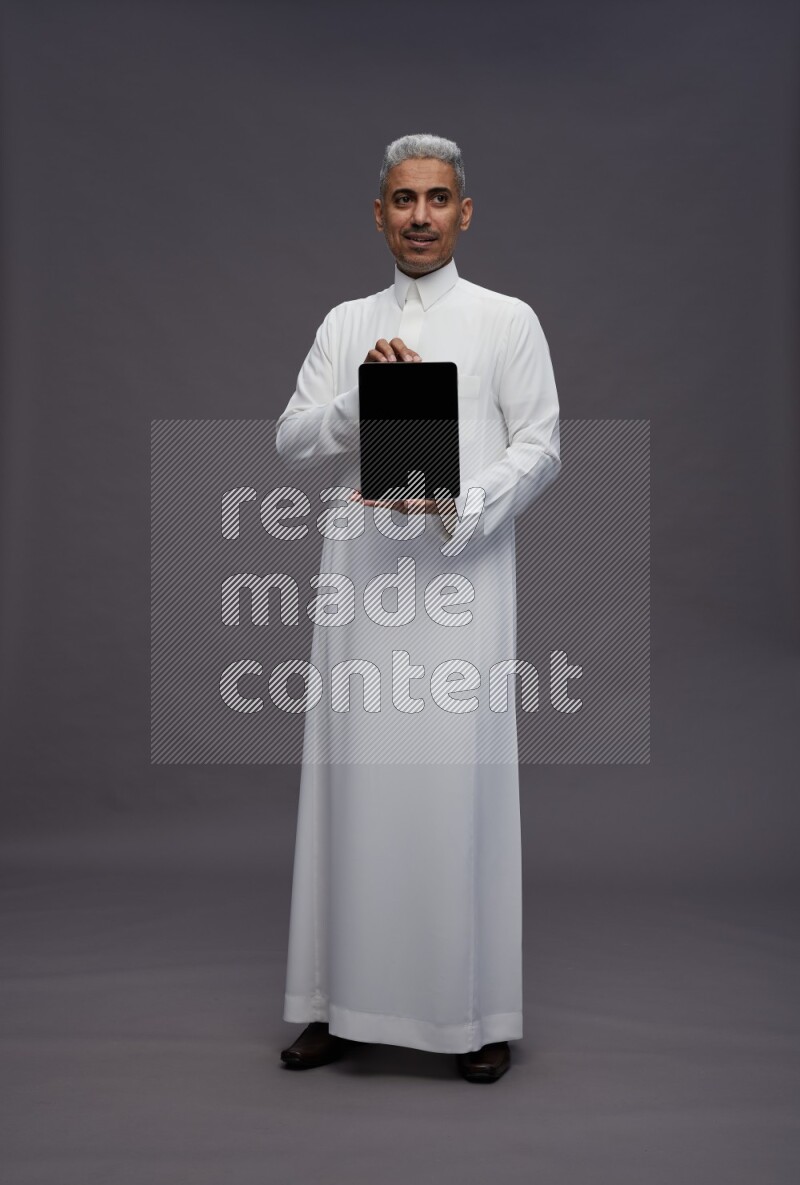 Saudi man wearing thob standing showing tablet to camera on gray background