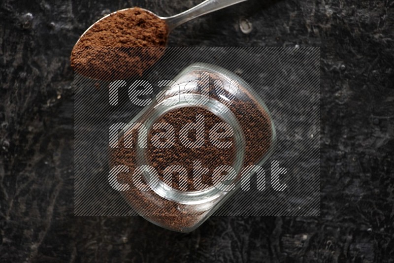 A glass spice jar and a metal spoon full of cloves powder on textured black flooring