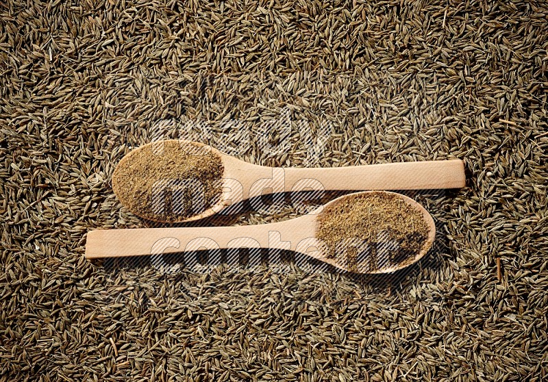 2 wooden spoons full of cumin powder on a cumin seeds background