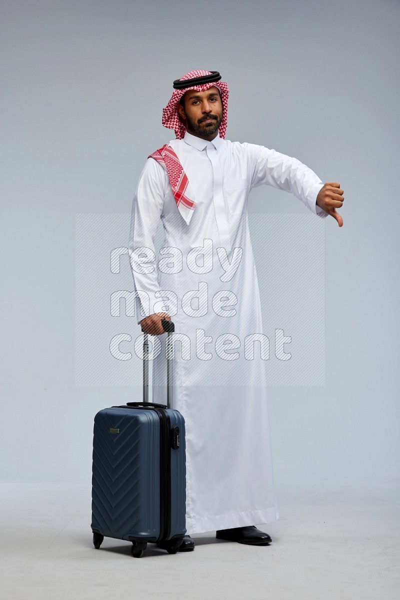 Saudi man wearing Thob and shomag standing holding Travel bag on Gray background