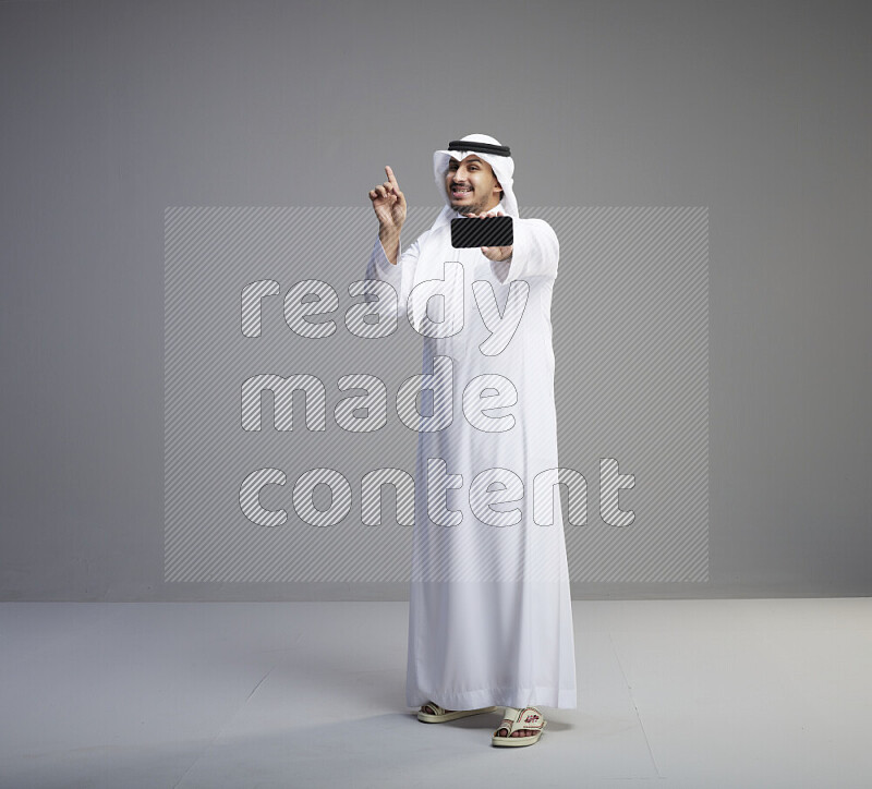 A Saudi man standing wearing thob and white shomag showing phone to camera on gray background