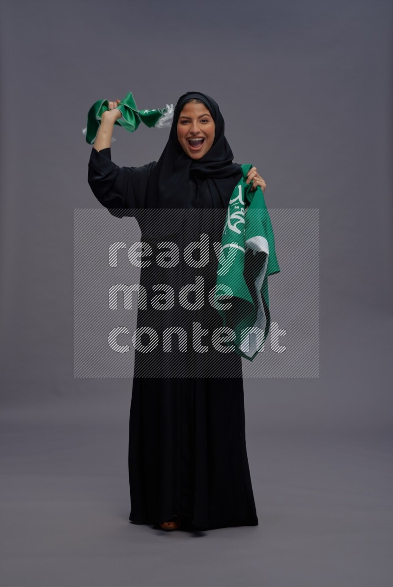 Saudi woman wearing Abaya standing holding Saudi flag on gray background
