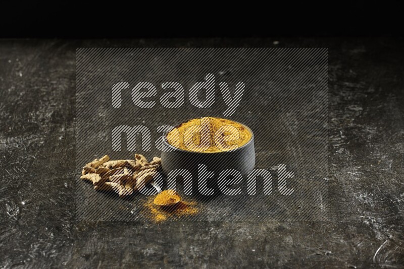 وعاء فخاري أسود وملعقة معدنية ممتلئة ببودرة الكركم مع أصابع الكركم المجففة على خلفية سوداء