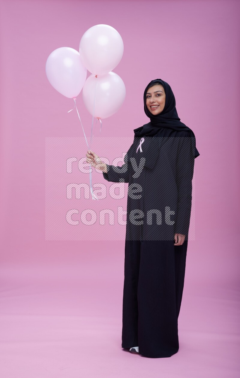 Saudi woman wearing pink ribbon on Abaya standing holding balloons on plain pink background