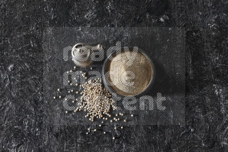 Black pottery bowl full of white pepper powder with pepper beads and metal pepper grinder on textured black flooring
