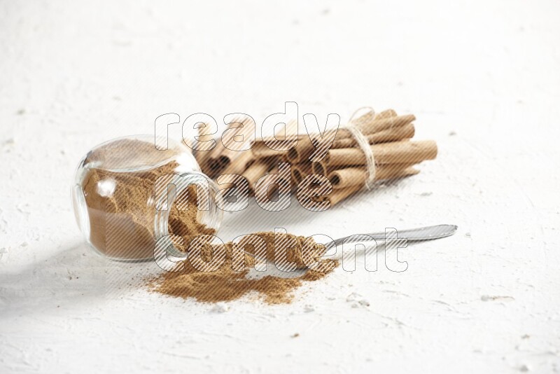 Flipped herbs glass jar full of cinnamon powder with a metal spoon full of powder and cinnamon sticks on a textured white background