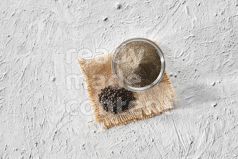 A glass bowl full of black pepper powder and black pepper beads on burlap fabric on textured white flooring