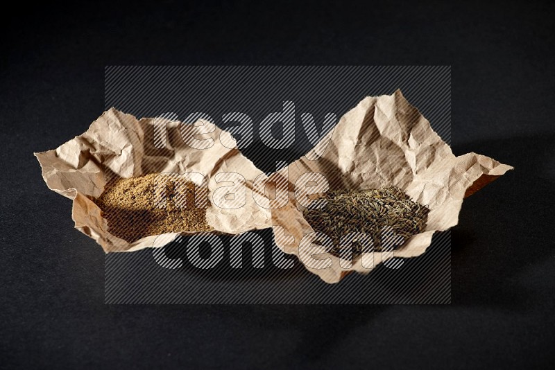 Cumin seeds and powder in 2 crumpled pieces of paper on black flooring