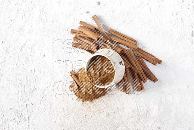 Ceramic beige bowl over filled with cinnamon powder and cinnamon sticks around the bowl on a textured white background in different angles