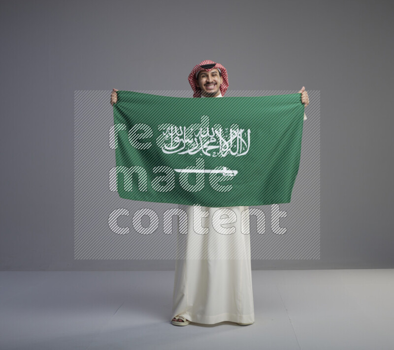 A saudi man standing wearing thob and red shomag holding big saudi flag on gray background