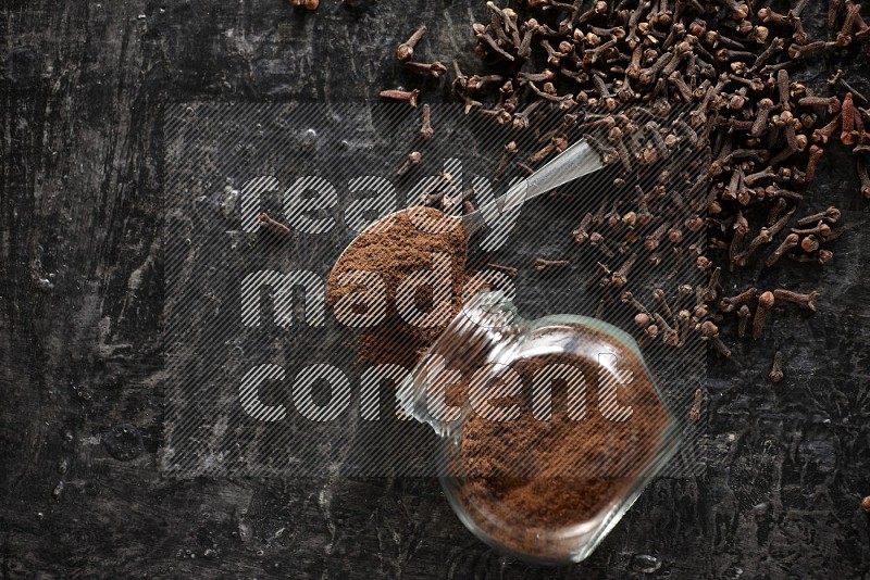 A flipped glass spice jar and a metal spoon full of cloves powder with cloves spread on textured black flooring